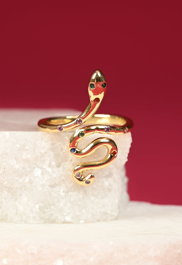 Gold snake-shaped ring with red accents on a white stone surface with a red background